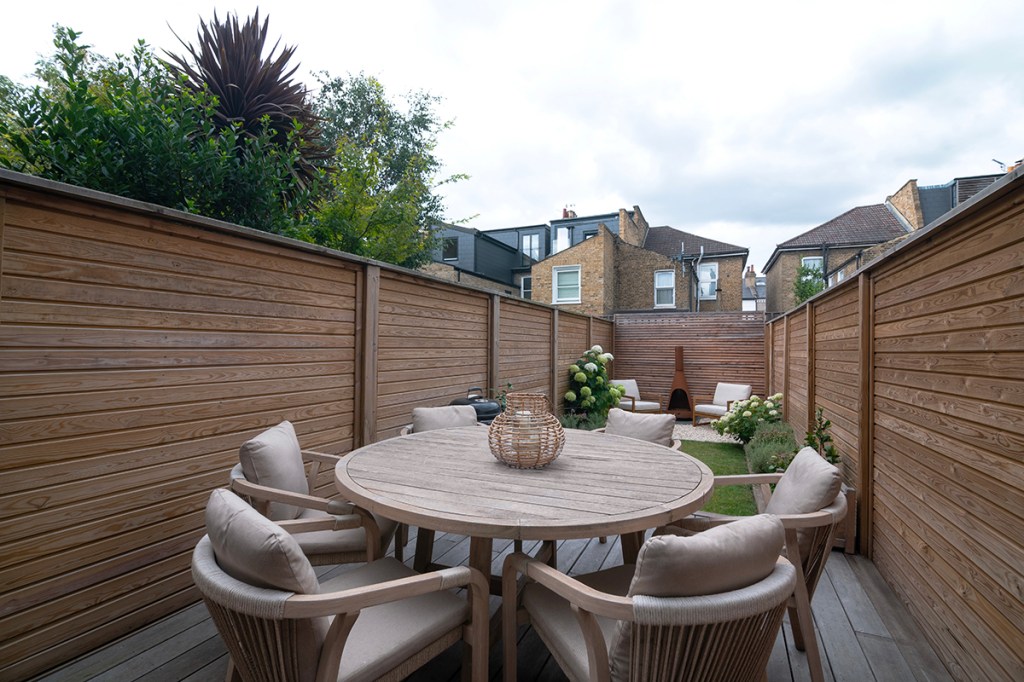 Landscaped garden in Balham, London featuring wooden decking, white stone flooring, and a rustic fireplace.