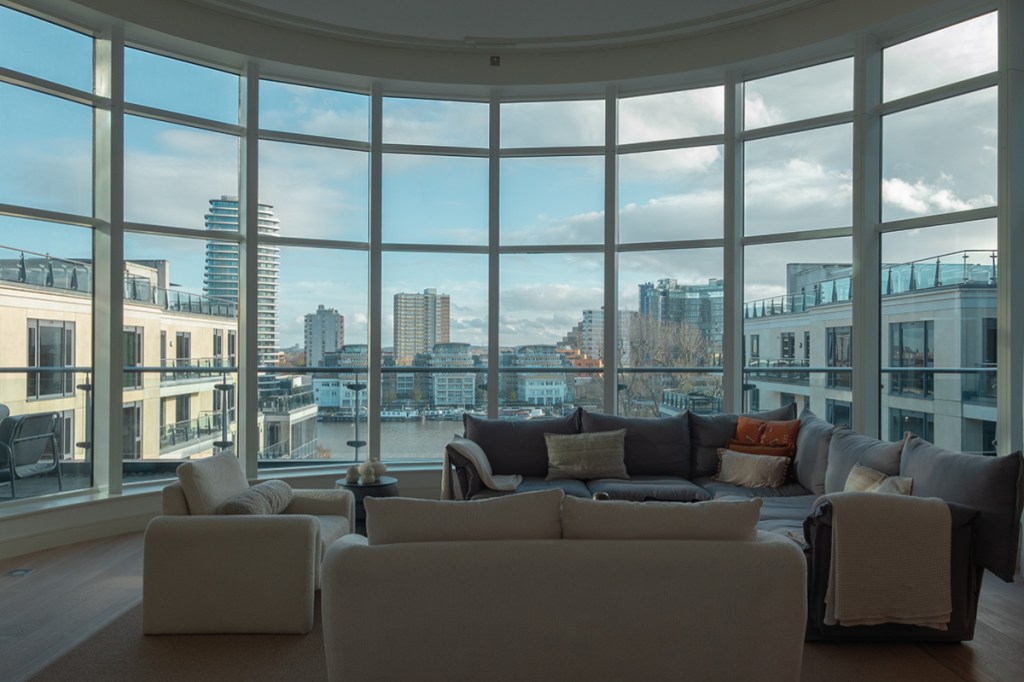 Top-floor penthouse living room in Chelsea, London with minimalist furniture and a large bay window framing the skyline.